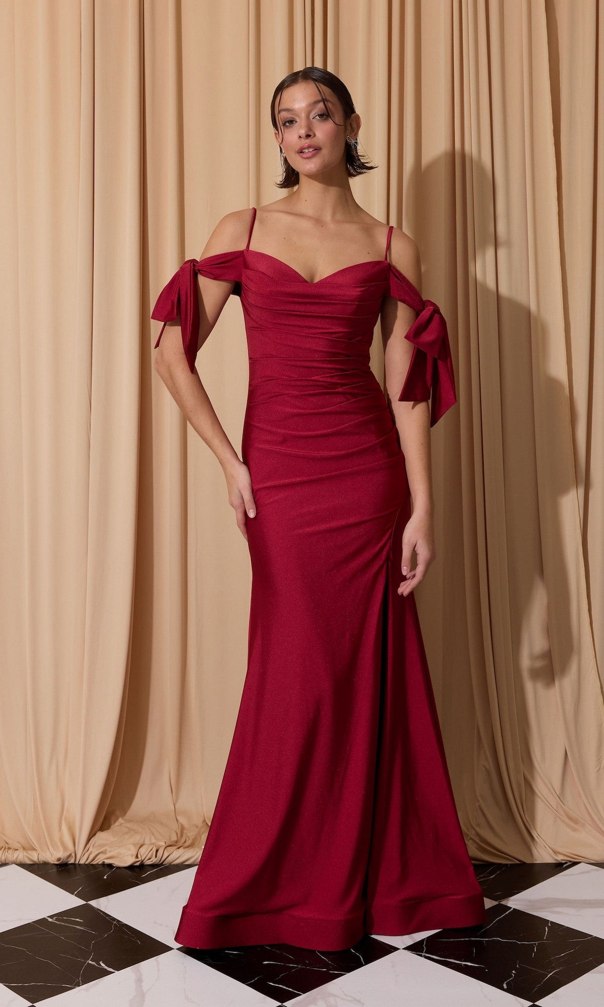 A woman stands before beige curtains wearing the Shoulder-Ties Long Jersey Prom Dress Eureka 9181 in burgundy, a floor-length design with ruched details and ribbon ties on the shoulders.